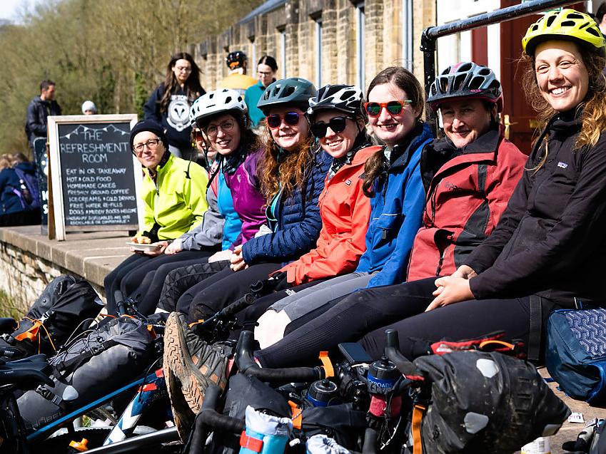 A group of women is sitting on a wall outside a café. They're all cyclists and are wearing cycling gear. You can see their bikes which are all heavily laden with luggage