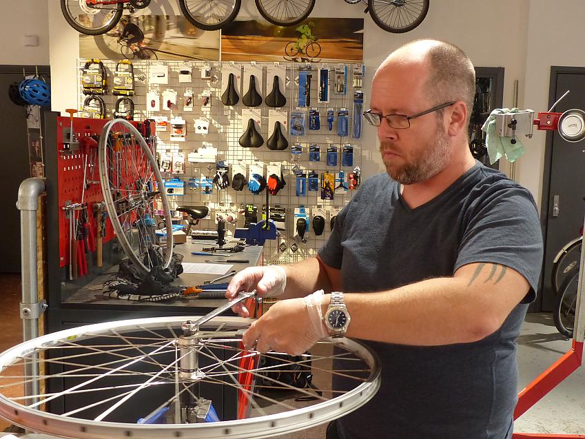 A man in a cycle workshop is working on a bicycle wheel. He has a beard and glasses. There are bike parts and accessories in the background