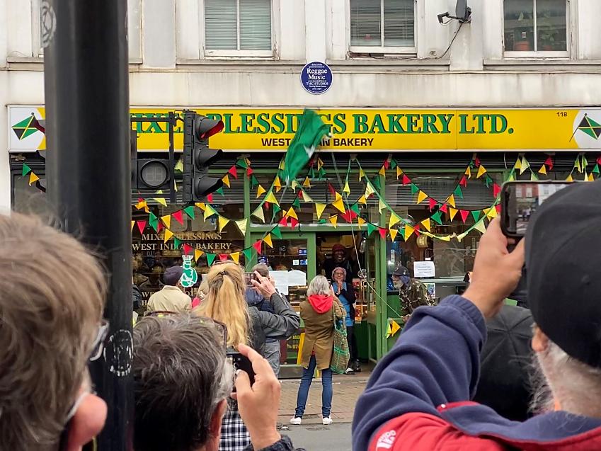 A celebration of the unveiling of a blue plaque on a West Indian bakery, which was previously a recording studio. Lots of people are taking photos and there is bunting all over the bakery in the colours of the Jamaican flag