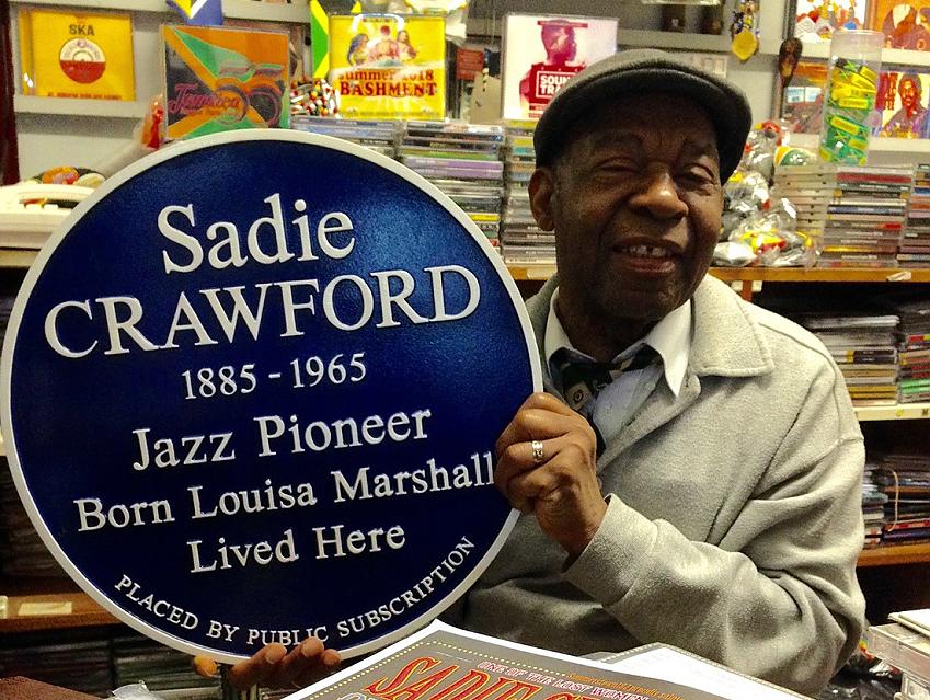 A shopkeeper is holding up a blue plaque recognising Sadie Crawford, a jazz performer. He is wearing a grey fleece and black flat cap. There are lots of CDs in the background.
