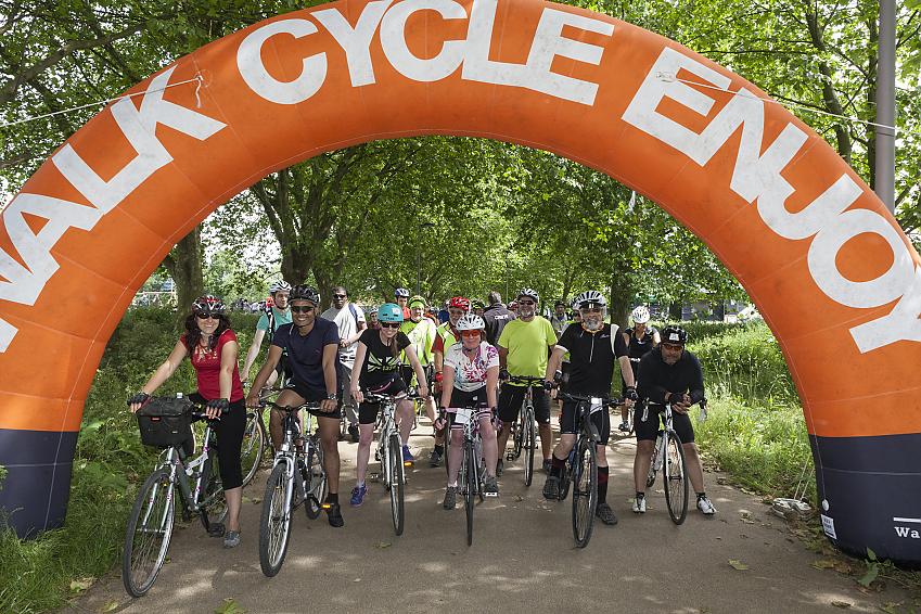 A group of cyclists is lined up underneath a big orange arch printed with the words 'Walk Cycle Enjoy'. They are about to set off on a ride