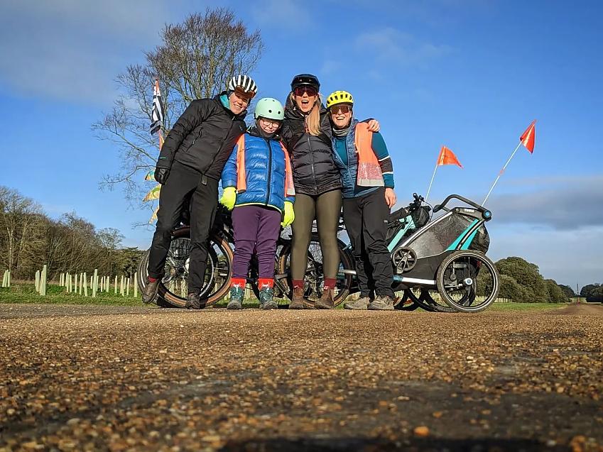 Four people (a woman, a young girl and two more women) are standing in front of a bike with a child trailer. They are all wearing warm cycling gear and smiling
