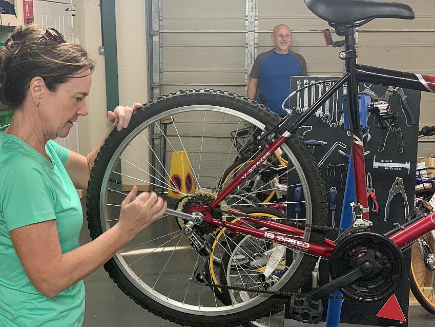 A woman in a green T-shirt is adjusting the gears on the back wheel of a bike. The bike is black and red and mounted on a stand so she can work on it. Behind the bike is a board with lots of bike tools on it.