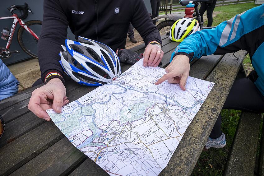 A close-up showing a paper map laid out on a pub table. One person is holding it while another is pointing at it. Both people are wearing cycling clothing and their helmets are on the table