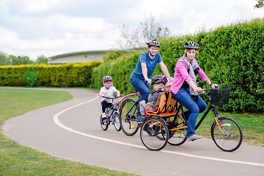 A family is cycling together. Mum is on a trike with two young children on the back, Dad has an older child on a trailer behind him