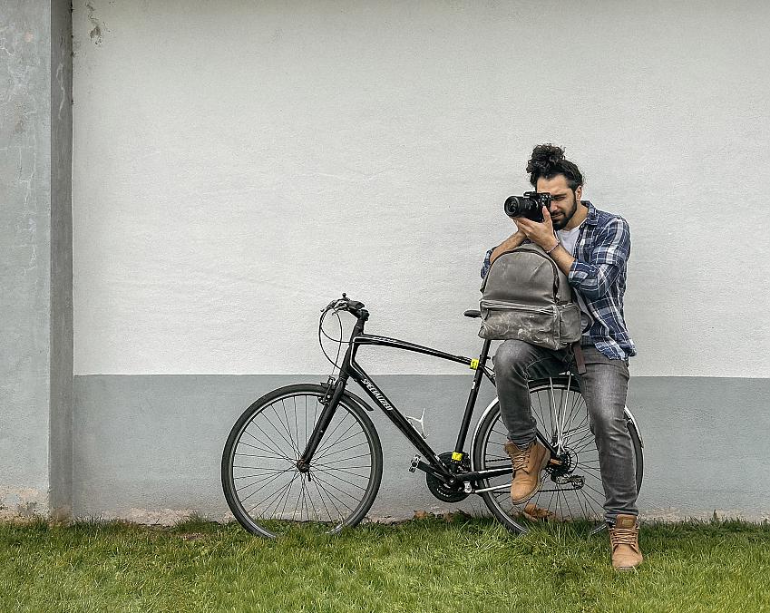 A man is perched on the rear rack of a black hybrid bike leaning against a white wall. He's wearing boots, black jeans and a check shirt. He has a rucksack on his lap and is resting his arms on it to hold up a camera which he's looking through