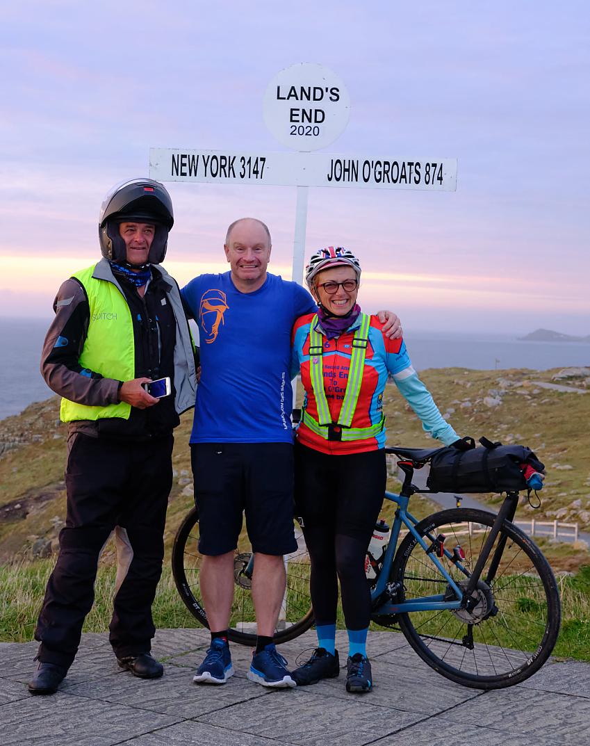 Two men and a woman are standing in front of the Land's End sign in Cornwall. The woman is wearing cycling gear and there is a blue bike behind her. One man is wearing shorts and T-shirt, the other is in motorbike gear and helmet