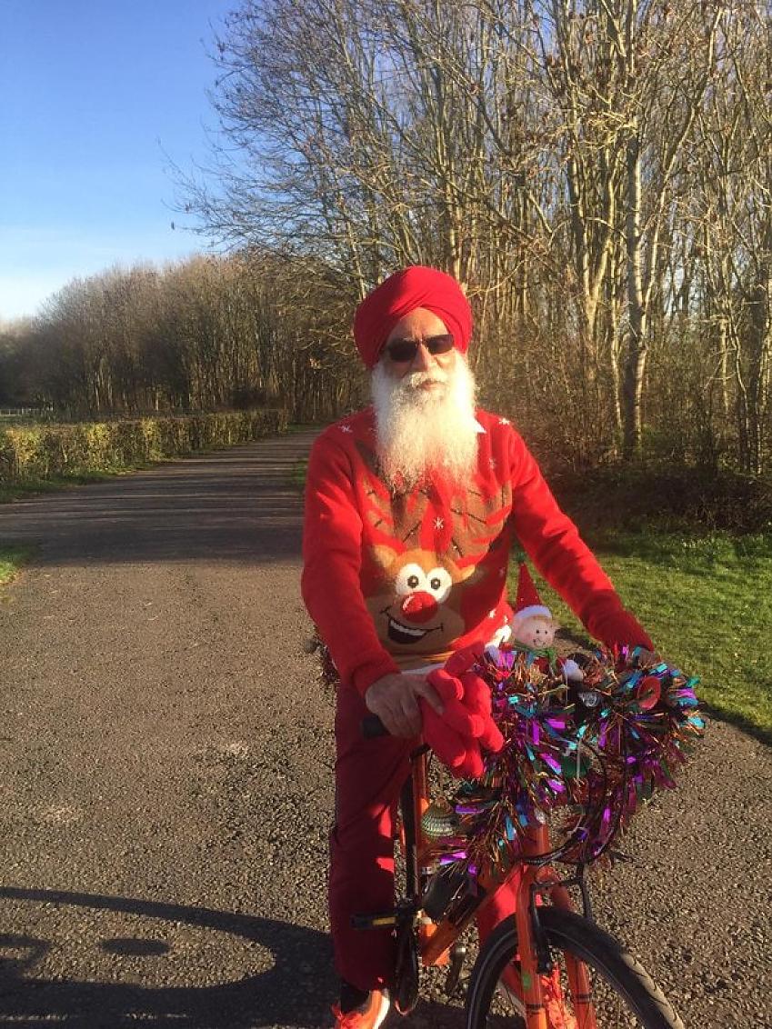 A male cyclist with beard and red turban wearing a reindeer jumper and bright red trousers; his bike is decorated with tinsel and Christmas elves