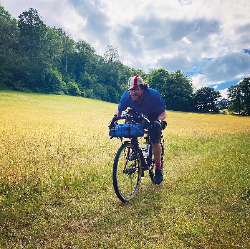 A man is riding a touring bike across a field. He's wearing the Pearl Izumi Men’s Rove Short Sleeve Shirt, shorts and a cap with the colours of the Italian flag.