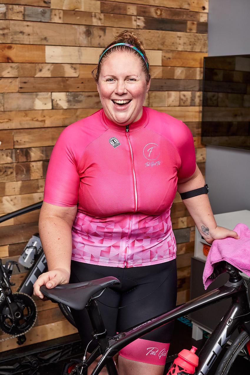 A woman stands with her bike. She is wearing a bright pink cycling jersey and black shorts with matching pink cuffs. She is smiling broadly