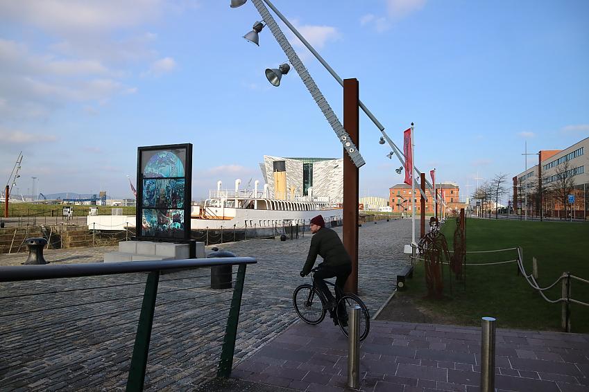 A man on a bicycle rides into a dockland area with a large ship visible in the background