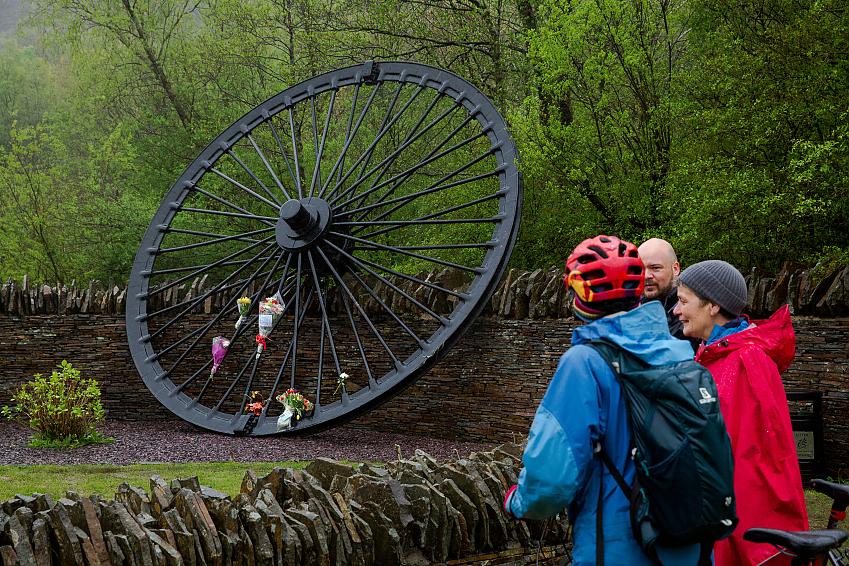 People stand beside a large black memorial wheel with flowers tied to it