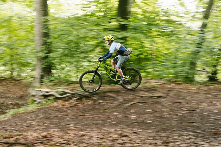 A man is cycling through a forest on a lime green mountain bike. He's performing a jump so flying through the air