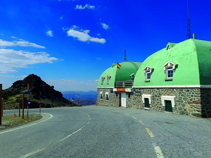 Green-topped stone buildings stand at the side of the road, a mountainous vista in the background