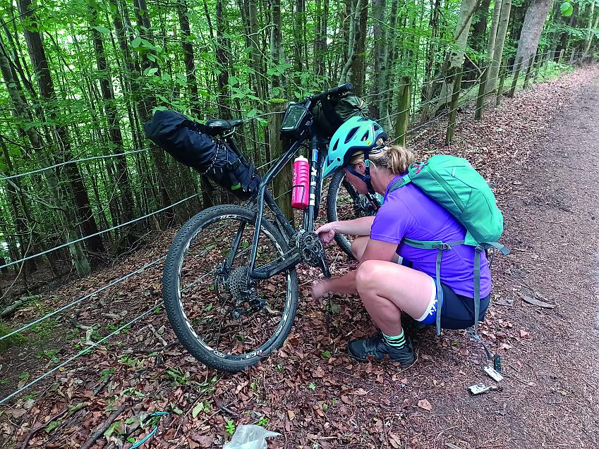 On a woodland path, a woman crouches down working on her bicycle