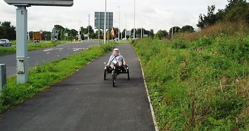 A woman on a tricycle is cycling along a wide cycle path, separated from the road by a grass verge