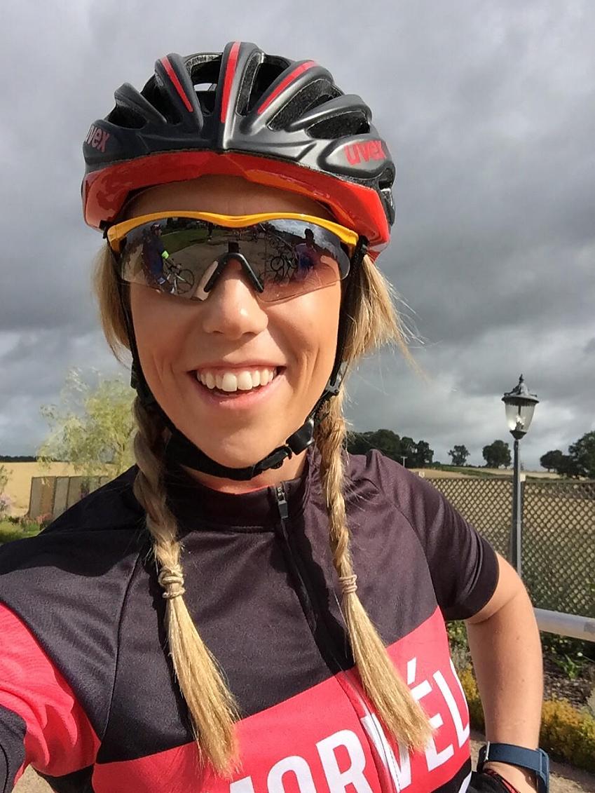 A woman in cycling gear is smiling at the camera. She has blonde plaits and is wearing a red and black helmet, sunglasses and a red and black cycling jersey