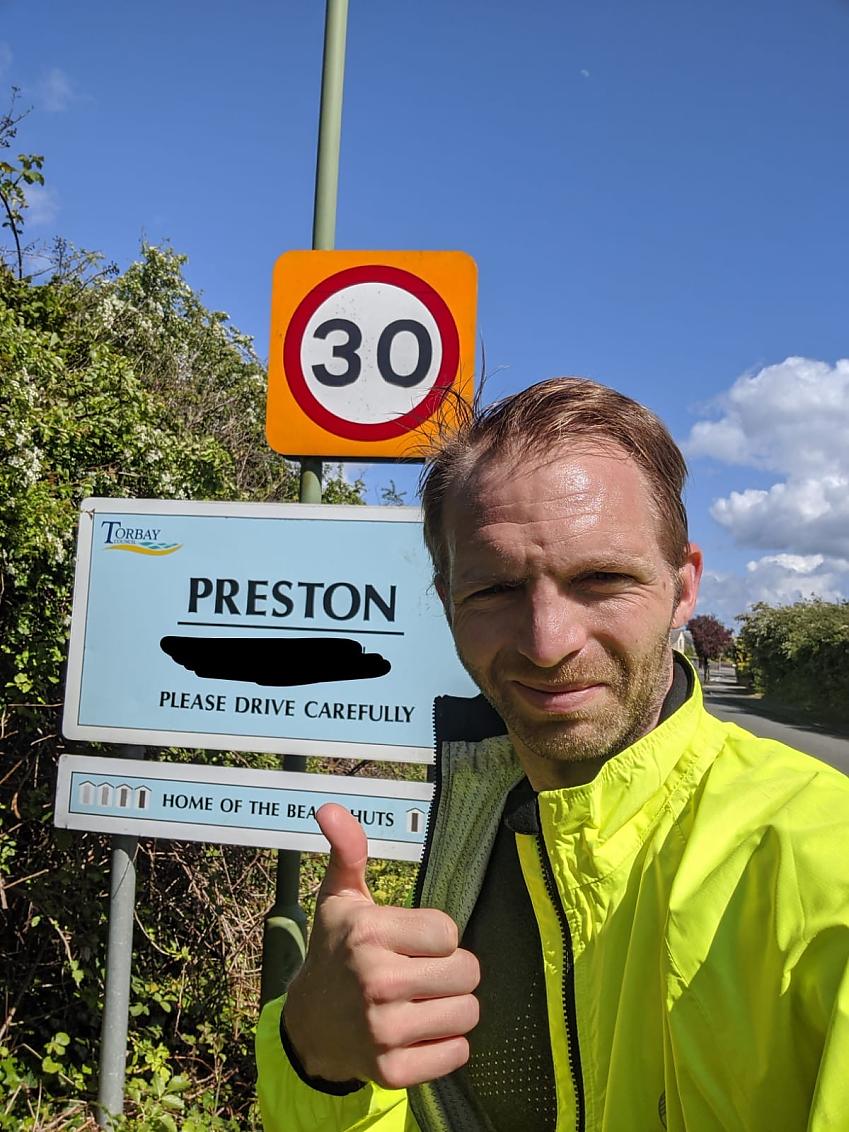 A male cyclist is standing in front of a sign for Preston in Devon. He is wearing a yellow hi-vis jackets and is giving a thumps-up