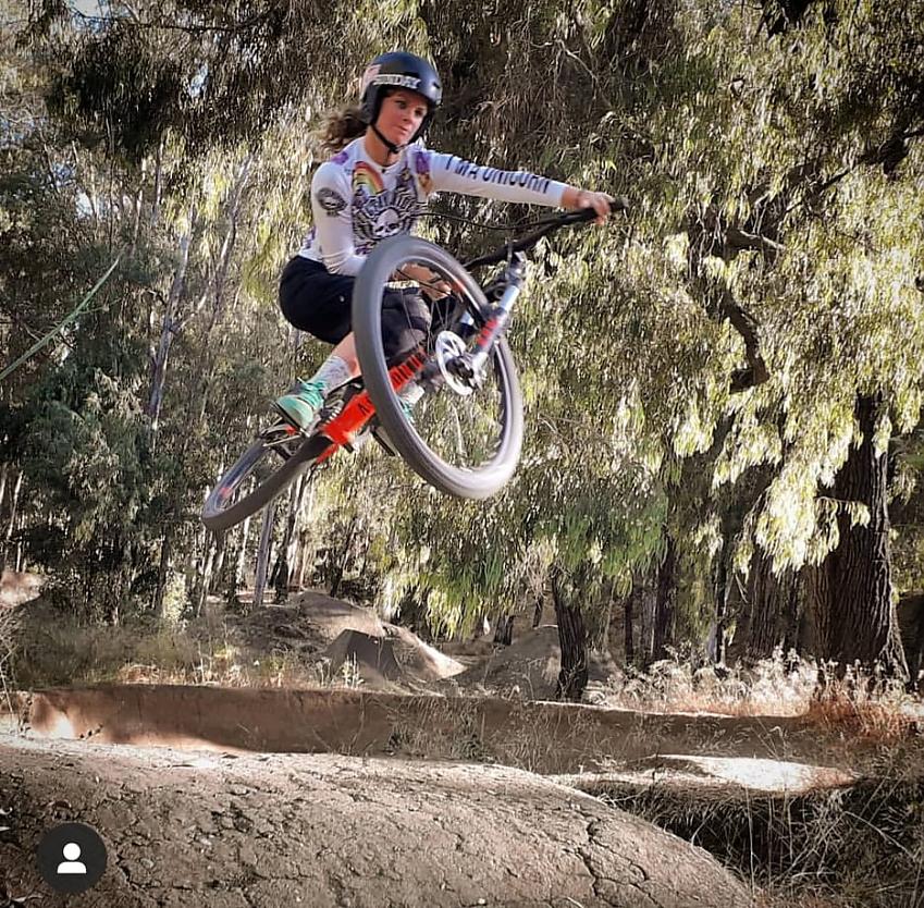 A woman is performing a jump on a mountain bike. She is in a forest setting. Her bike is orange and she's wearing black three-quarter-length trousers and a white jersey with a skull and rainbow design