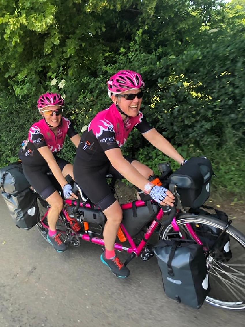 Two smiling ladies cycle towards us on a bright pink tandem; they're wearing matching pink cycle clothing