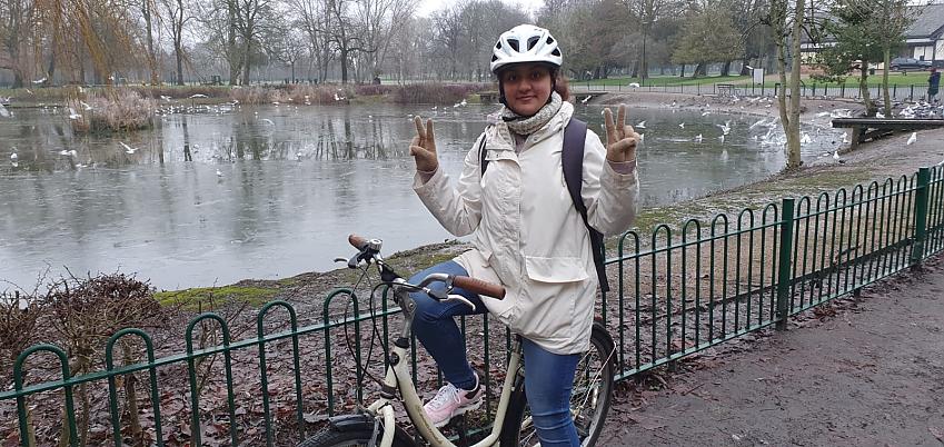 A young woman is on a bike. She is posing in front of a frozen lake. She is wearing blue jeans and a white winter jacket. She is smiling and holding her hands up making peace signs