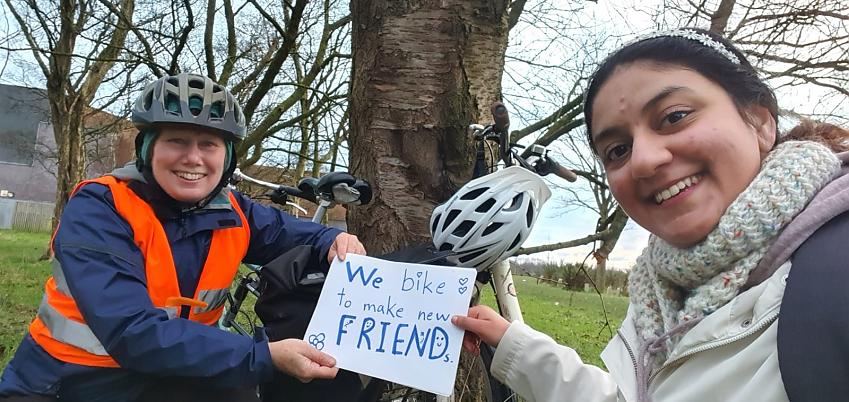 Two women are standing in front of a tree with a white bike leaning against it. They are holding a sign that reads 'We bike to make new friends'. It's winter and they are wearing warm clothing. They're both smiling happily.