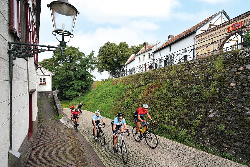 Cyclists ride along a cobbled street flanked by houses either side