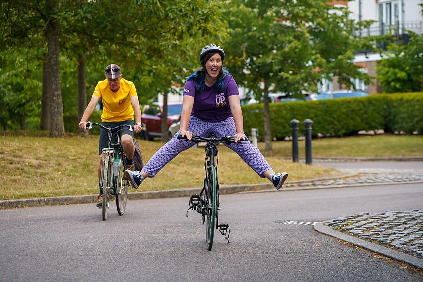 Two people riding cycles. The woman is in front, she is wearing bright purple, has her legs stretched out in front of her and a big smile; a man is cycling behind her, he is wearing a bright yellow T-shirt