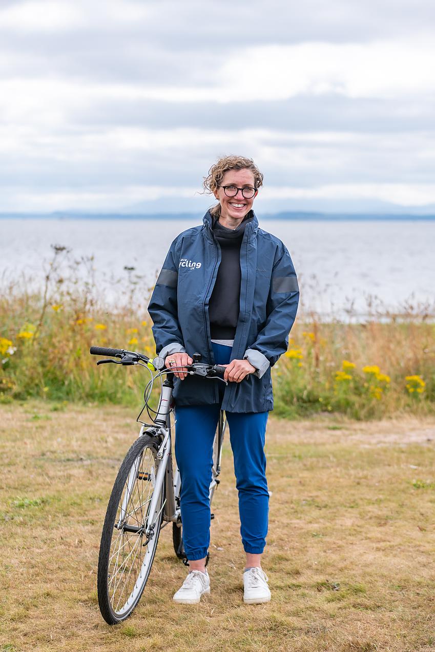 A woman is standing with a silver bike. She's wearing a blue Cycling UK jacket and blue trousers. 