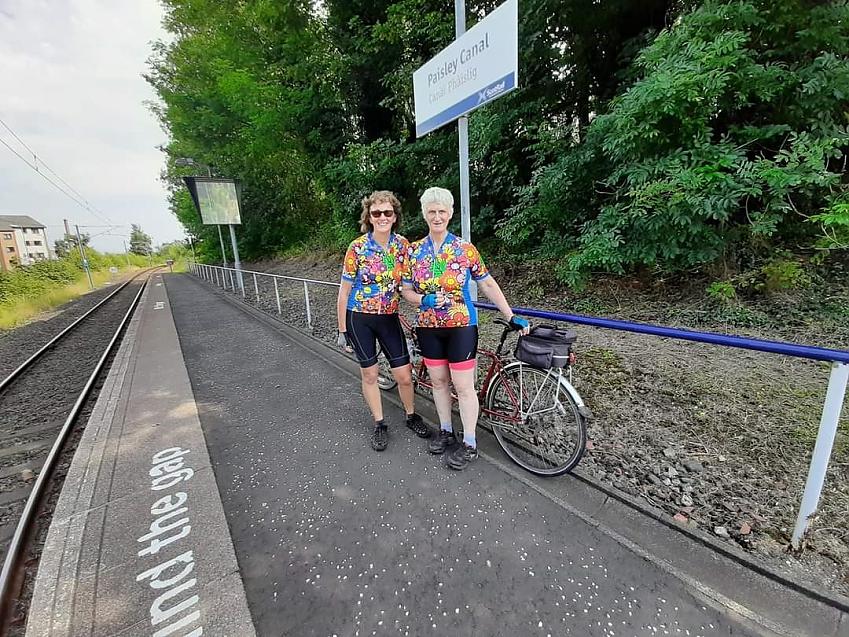 Two women are standing side by side on a railway platform. They are wearing matching flower-print cycling jerseys. Behind them is a red tandem