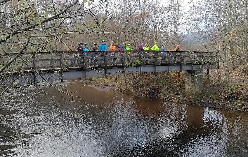 A group of cyclists are all standing on a bridge over a stream. They are all wearing bright jackets and a couple are waving at the camera. It looks like it's autumn and the trees are mostly bare