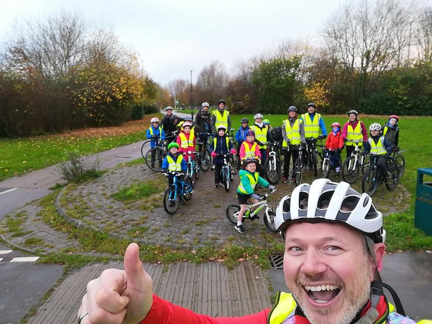A man has taken a selfie showing his head and one hand doing a 'thumbs up' in the foreground. He's wearing a cycle helmet. In the background is a group of kids and parents on their bikes