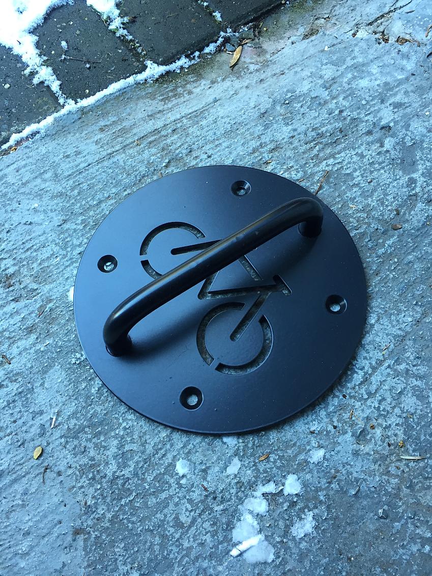 A black bike anchor has been placed on the ground, which looks like concrete. It hasn't been bolted in
