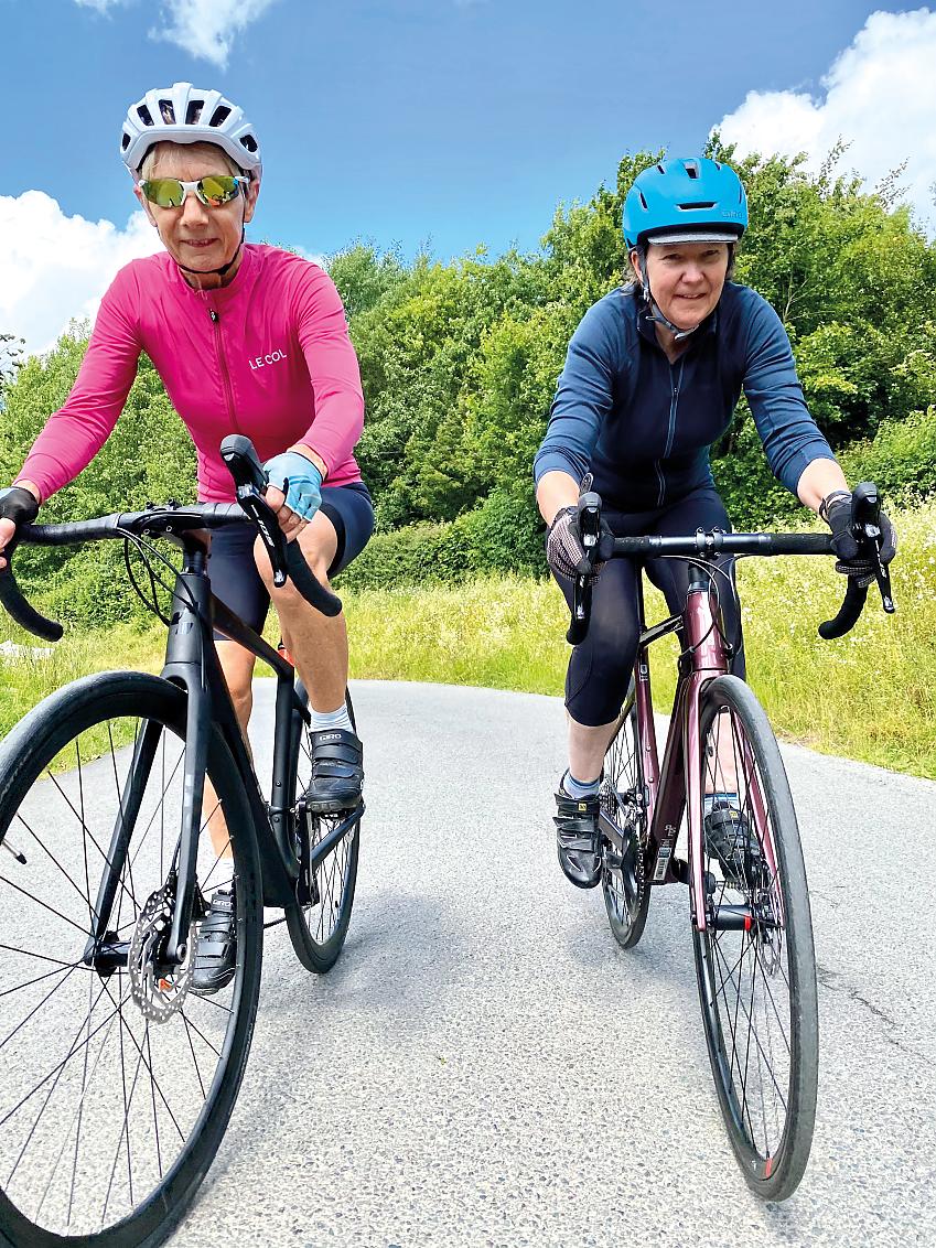 Two women are cycling towards the camera on road bikes. On the left is a black bike with a rider in pink and black, on the right is a purple bike with a rider in blue and black.