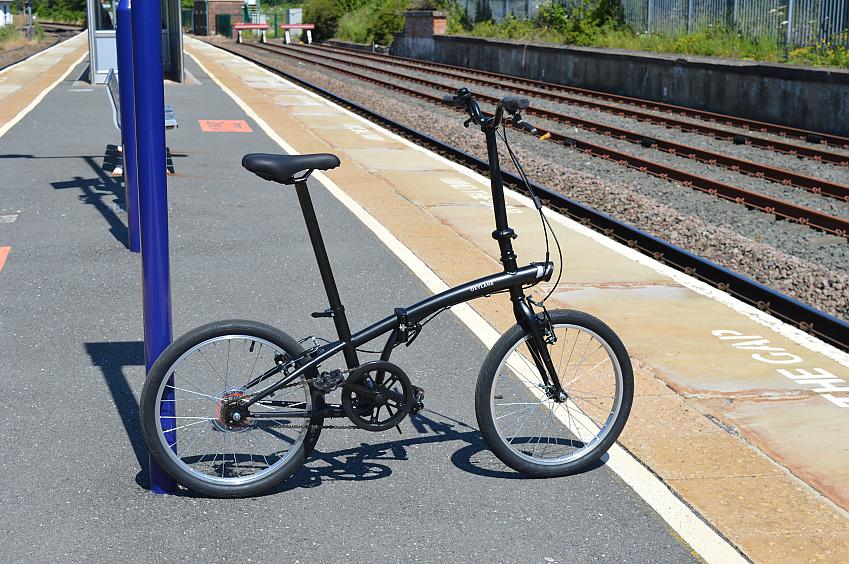 A B'twin black folding bike is propped up against a pole on a railway station platform