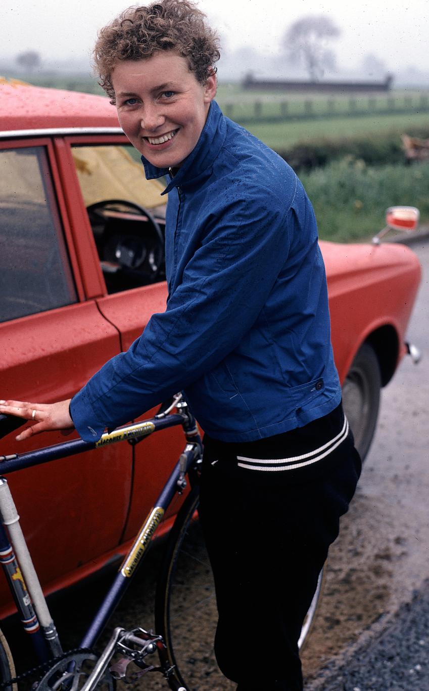 A woman, Beryl Burton, with her bike, which is blue, next to an orange car