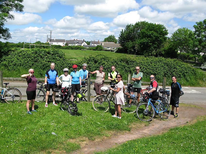A group of cyclists are standing on the grass with their bikes. There is a variety of bikes and some people are wearing cycling gear, others normal clothes