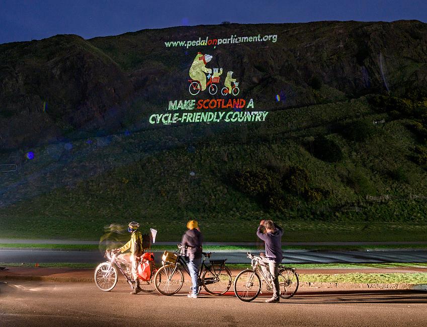 The message 'Make Scotland a cycle-friendly country' is projected onto a hillside in Edinburgh. Three people on bikes are in the foreground looking up at the projection
