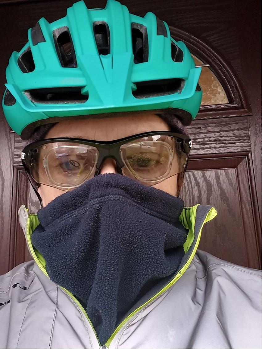 A woman is wearing a turquoise cycling helmet and silver jacket. She has cycling sunglasses on and a blue face covering over her mouth and nose