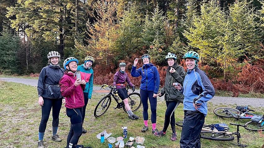 A group of women with their bikes have stopped to have some food. A selection of cheeses and crackers are laid out on the ground in front of them