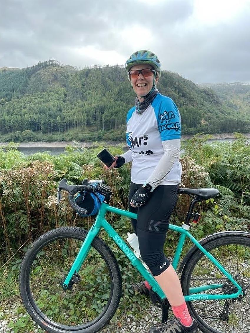 A woman is standing with her Sonder gravel bike. She is wearing black cycling shorts and a Mrs Bump cycling jersey. In the background are mountains and a lake