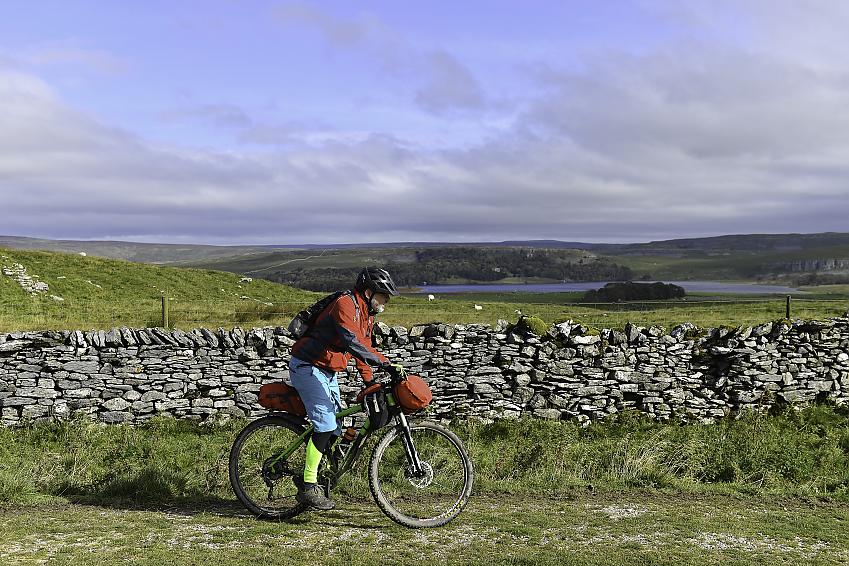 A man on a bike loaded with bags is cycling through the Yorkshire Dales countryside, past a dry stone wall. He's wearing an orange jacket, blue shorts and black leggings. There's a lake in the background