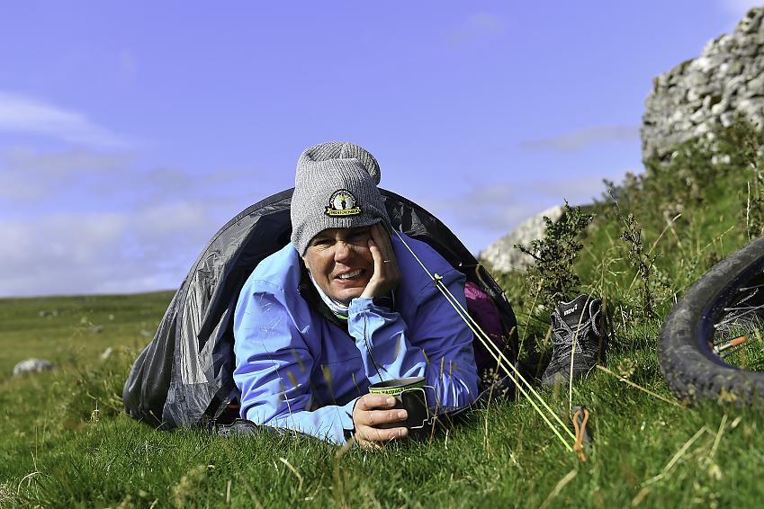 A woman is holding a travel cup of coffee while lying in a bivvy bag. It's morning and she's slept there overnight. She's wearing a blue jacket and smiling. One walking shoe and a bit of a bike wheel can just be seen in shot