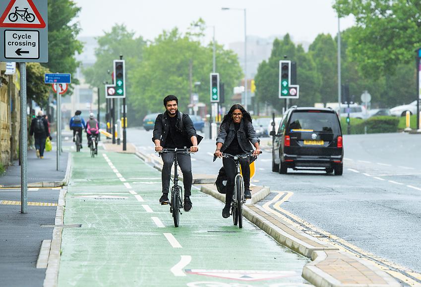 Multi-modal travel with a pavement, cycle path and road. In the foreground, two people are cycling together on the cycle path. They are wearing normal clothes and have bags. They are smiling. In the background, more people are cycling, walking and driving