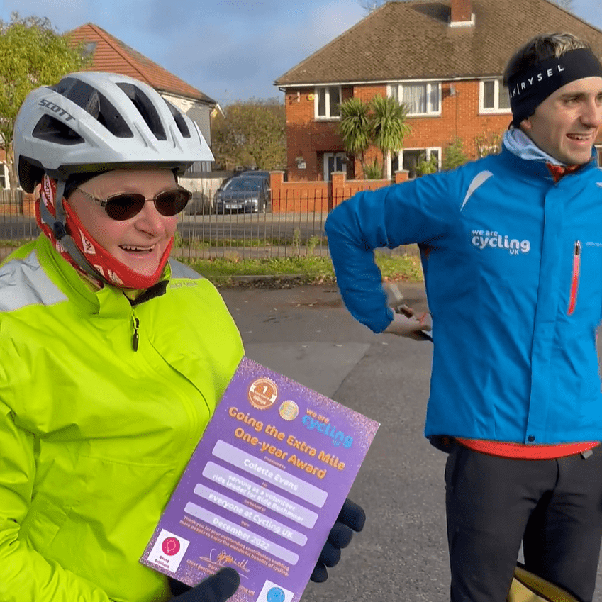 Two people in cycling gear. At the front is a woman wearing a hi-vis waterproof cycling jacket. She is holding a purple award. In the background is a man in a blue We are Cycling UK jacket