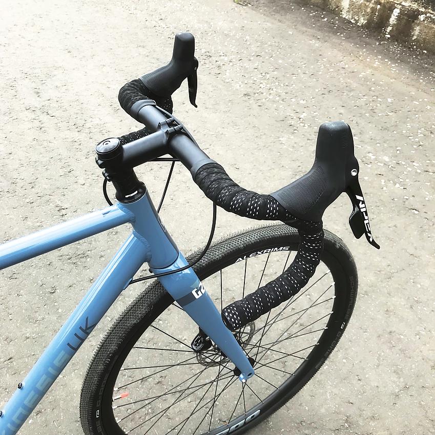 Front end of the Kinesis G2, a light blue gravel bike, showing the handlebar, brakes, gear levers and hoods, front wheel