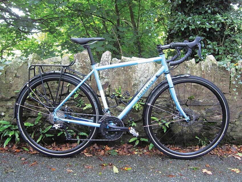 Ridgeback Panorama, a light blue touring bike with drop bar, rear rack and mudguards, leaning against a wall