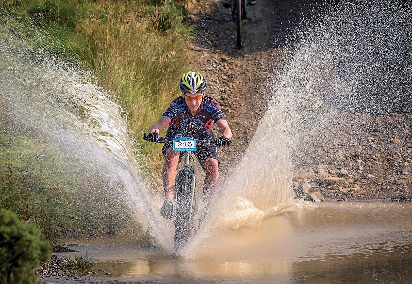 A man is riding a mountain bike through water. There is water spraying all around him. He has a number (216) on the front of the bike, indicating that he's taking part in a sporting event