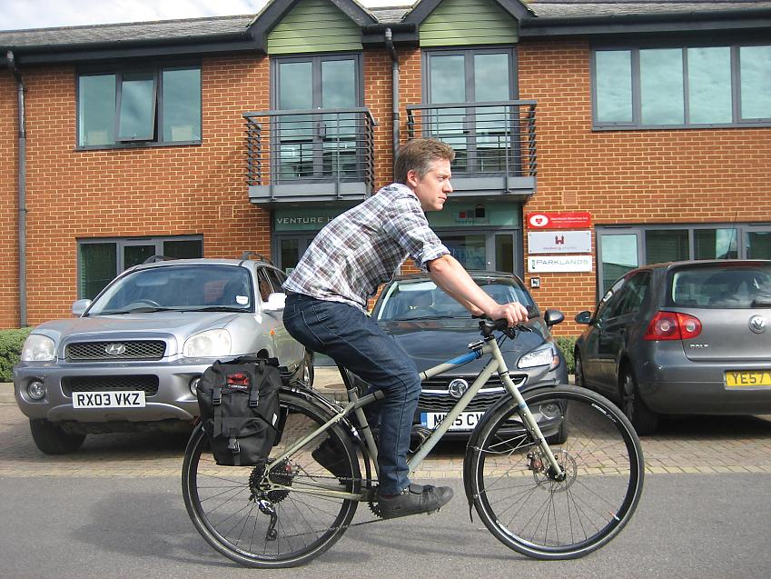 A man is riding a bike past some cars parked in front of an office. He is wearing normal clothes and the bike has a small pannier on the rear rack