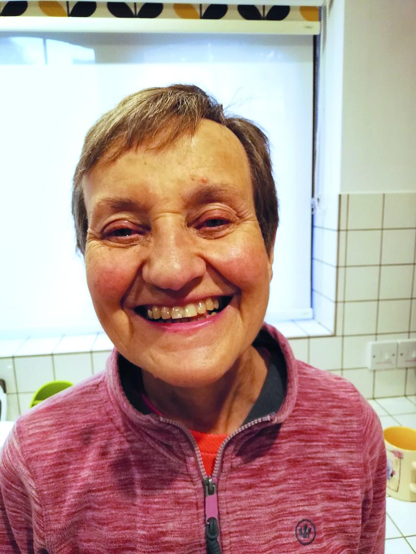An older woman is seen smiling at the camera. She is wearing a pink fleece and is standing in a kitchen. She has short cropped hair and a warm broad smile.